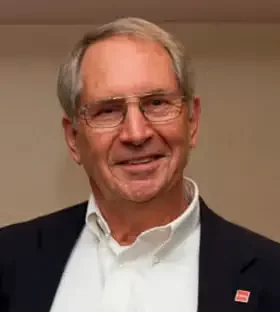 A smiling middle-aged man with gray hair and glasses, wearing a white collared shirt and a dark jacket, standing indoors.