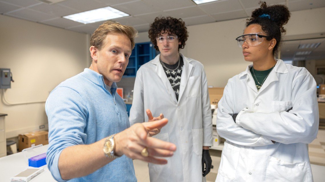 A man explaining something to two young women in a laboratory, all wearing lab coats and safety glasses.